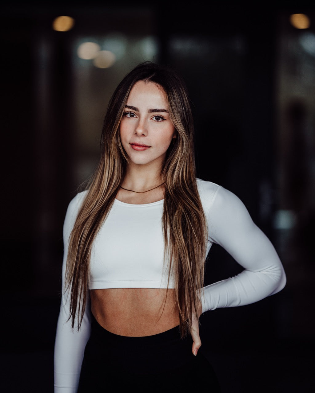 Woman wearing a white long-sleeve crop top and black pants against a dark background